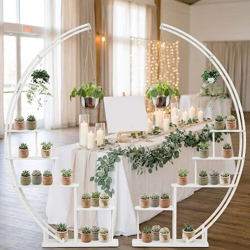 Two white half-moon tiered stands holding mini succulent wedding favors beside a greenery-draped reception table.